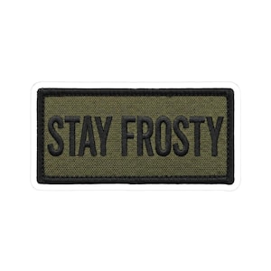 May include: A rectangular patch with the words "STAY FROSTY" embroidered in black on an olive green background. The patch has a black border and is likely designed to be sewn or attached to clothing or gear.
