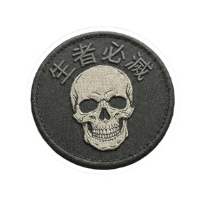 May include: A round, dark gray patch with a skull design in the center. The skull is light gray and detailed, with visible teeth and eye sockets. Above the skull are Japanese characters. The patch has a stitched border.