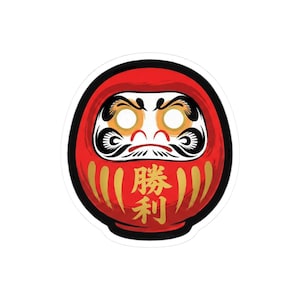 May include: A red Daruma doll sticker with black outlines and gold accents. The doll has a detailed face with white eyes, black eyebrows, and a small red mouth. The Japanese characters for "victory" are written vertically in gold.