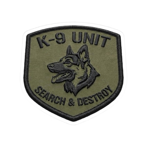 May include: An olive green patch with a black border and the words "K-9 UNIT" at the top. Below is a black embroidered image of a dog's head, and the words "SEARCH & DESTROY" are at the bottom.