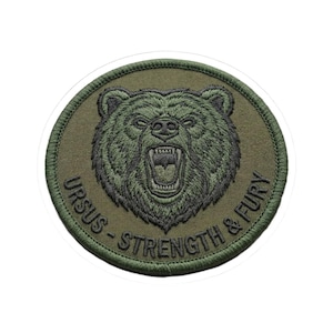 May include: Round olive green patch featuring a roaring bear's head with open mouth and visible teeth. The embroidered text around the bear reads "URSUS - STRENGTH & FURY" in a dark green color. The patch has a stitched border.
