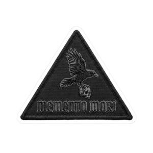 May include: Black triangular patch featuring a raven perched atop a skull. The raven has outstretched wings. The words "MEMENTO MORI" are embroidered in a gothic font below the skull. The patch has a white border.