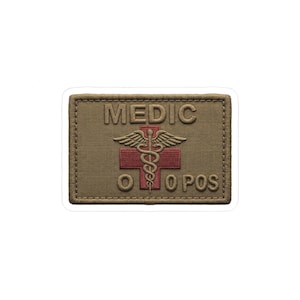 May include: A rectangular patch with the word "MEDIC" at the top. Below is a red cross with a caduceus symbol. The bottom of the patch reads "O O POS". The patch is tan with a stitched border.
