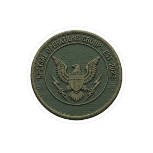 May include: A circular patch with a green background and a white border. The patch features a detailed eagle emblem with outstretched wings, a shield, and lightning bolts. The text "SPECIAL OPERATIONS GROUP - EST. 2023" is in a circular pattern around the eagle.