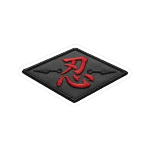 May include: Black and red embroidered patch in a diamond shape. The patch features a red Japanese Kanji symbol and two black kunai knives. The patch has a white border and is likely an iron-on or sew-on patch.