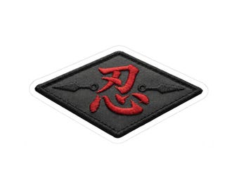May include: Black and red embroidered patch in a diamond shape. The patch features a red Japanese Kanji symbol and two black kunai knives. The patch has a white border and is likely an iron-on or sew-on patch.