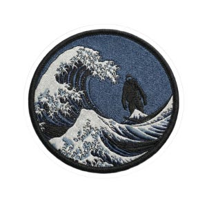 May include: Round patch featuring a stylized depiction of a large wave with a black silhouette of a figure standing on a smaller wave. The patch has a blue background and a black border. The wave is white and blue.