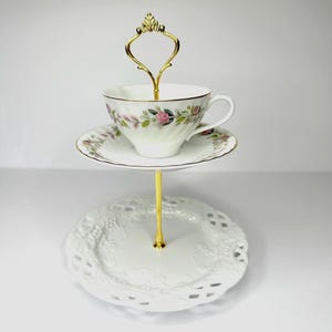 May include: A two-tiered serving stand with a gold handle and support. The top tier holds a teacup and saucer with a floral pattern. The bottom tier is a white, lacy plate. The teacup and saucer have gold trim.