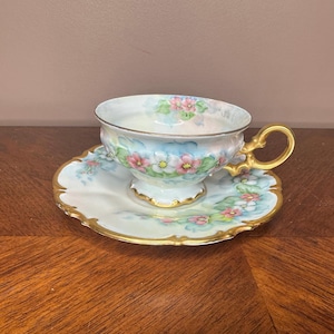 May include: A vintage teacup and saucer set. The cup and saucer are white with a light blue background and pink floral design. The edges are trimmed with gold. The handle of the cup is gold.