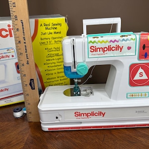 May include: A white and teal Simplicity First Fashions sewing machine with red accents. The machine has a handle, a needle, and a red and white safety symbol. A box and a ruler are in the background.