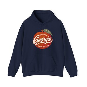 May include: A navy blue hooded sweatshirt featuring a graphic of a peach with a green leaf. The text on the peach reads Sweet as a Georgia Peach Est. 1788 in white and cream lettering. The hoodie has long sleeves and a front kangaroo pocket.