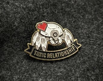 Op de afbeelding: Een wit en grijs robot spin emaille pin met een rood hart en de woorden "TOXIC RELATIONSHIP" op een zwarte banner met gouden rand. De pin is geplaatst tegen een donkergrijze, getextureerde achtergrond.
