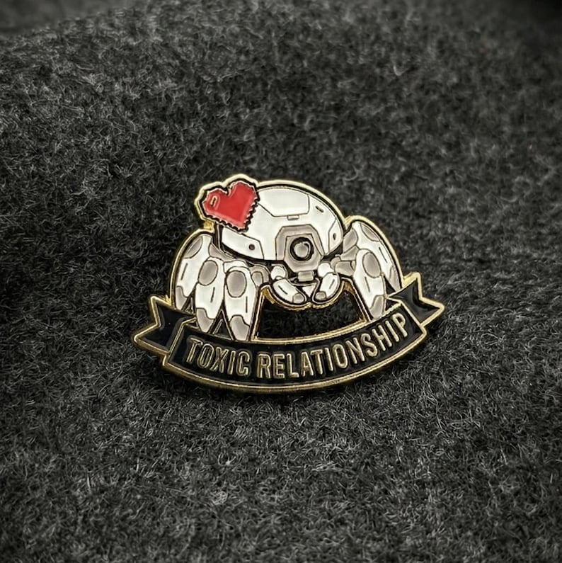 Op de afbeelding: Een wit en grijs robot spin emaille pin met een rood hart en de woorden "TOXIC RELATIONSHIP" op een zwarte banner met gouden rand. De pin is geplaatst tegen een donkergrijze, getextureerde achtergrond.
