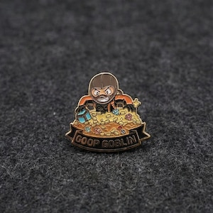 Op de afbeelding: Een goudkleurige emaille pin met een cartoon goblin karakter in een oranje en bruine outfit, die opduikt uit een stapel gouden munten en juwelen. De pin heeft de tekst "GOOP GOBLIN" op een banner.