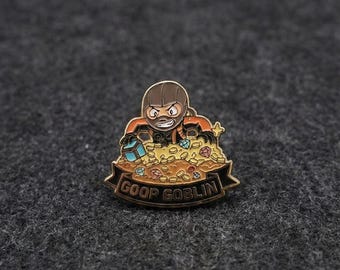 Op de afbeelding: Een goudkleurige emaille pin met een cartoon goblin karakter in een oranje en bruine outfit, die opduikt uit een stapel gouden munten en juwelen. De pin heeft de tekst "GOOP GOBLIN" op een banner.