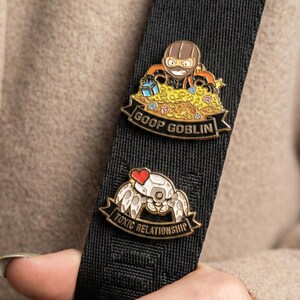 Op de afbeelding: Twee emaille pins op een zwarte band. De bovenste pin toont een cartoon goblin in een masker omringd door schatten, met de tekst "GOOP GOBLIN". De onderste pin heeft een robotkrab met een hart en de tekst "TOXIC RELATIONSHIP".