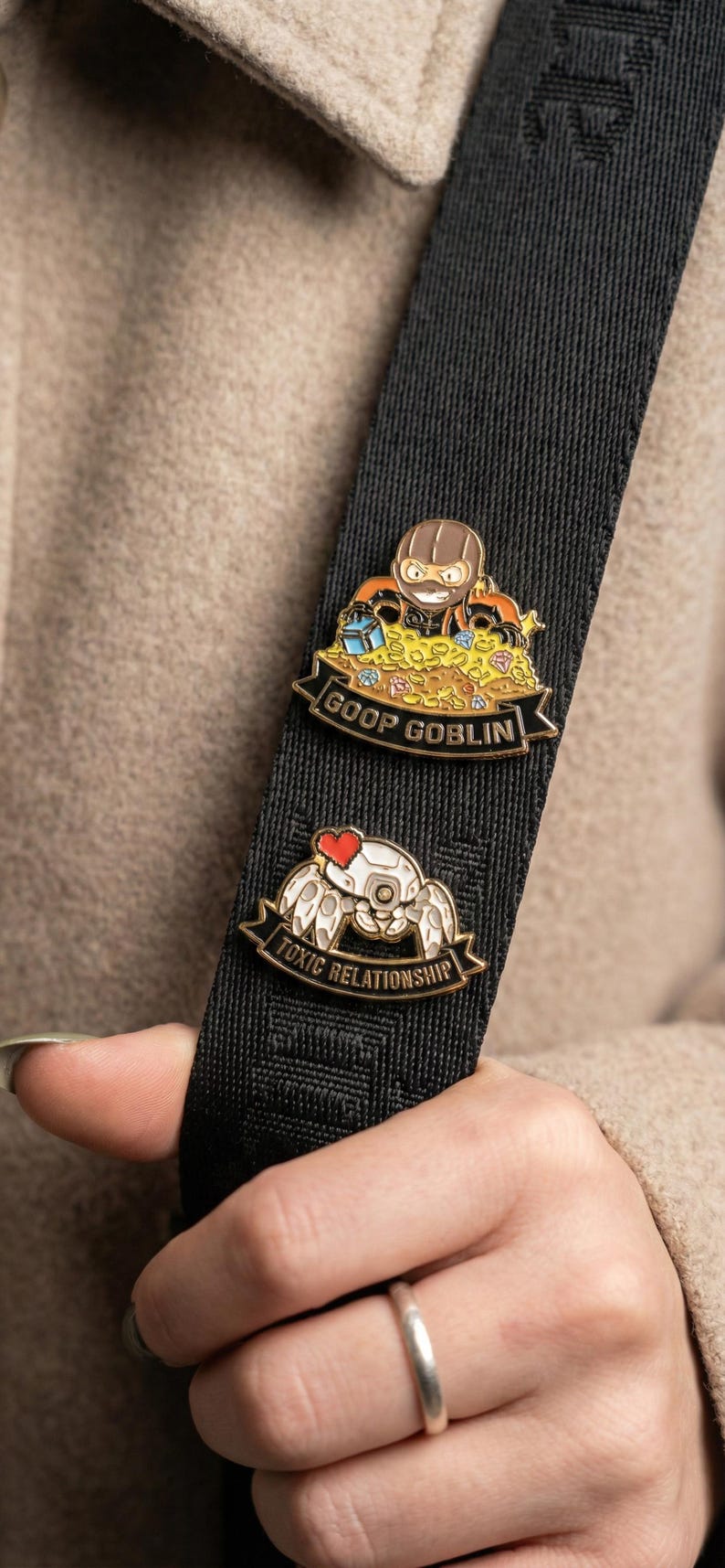 Op de afbeelding: Twee emaille pins op een zwarte band. De bovenste pin toont een cartoon goblin in een masker omringd door schatten, met de tekst "GOOP GOBLIN". De onderste pin heeft een robotkrab met een hart en de tekst "TOXIC RELATIONSHIP".