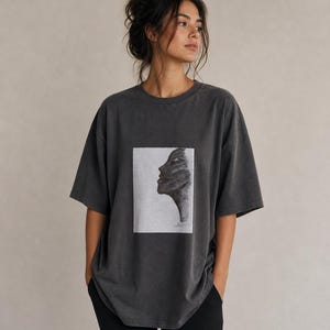Op de afbeelding: Donkergrijs t-shirt met een zwart-wit portret van een gezicht op de voorkant. Het shirt heeft een ronde hals en korte mouwen. De stof lijkt zacht en licht versleten, wat het een vintage uitstraling geeft. Het model draagt een zwarte broek.