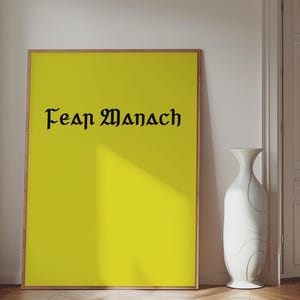 May include: A rectangular framed print featuring a solid bright yellow background with the black text "FEAR MANACH" written in a stylized gothic font. The frame is thin and light brown, positioned vertically against a plain white wall.