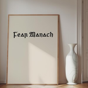 Puede incluir: Una impresión rectangular enmarcada que presenta el texto negro "Fear Manach" en una fuente gótica estilizada centrada sobre un fondo de color crema. El marco es de madera de tono claro y la obra se muestra en orientación vertical.