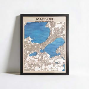 Könnte beinhalten: Gerahmte Holzkarte von Madison, Wisconsin, mit blauen Wasserelementen und detaillierten Straßen. Der Name der Stadt "MADISON" steht oben, darunter der Text "Custom text here: Amy & Jason 2021". Die Karte ist in einem schwarzen Rahmen.