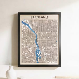 May include: Framed wooden map of Portland, Oregon, with a black frame. The map features a detailed street layout and a blue river running through the city. Text at the top reads "PORTLAND" and "Custom text here: Amy & Jason 2021".