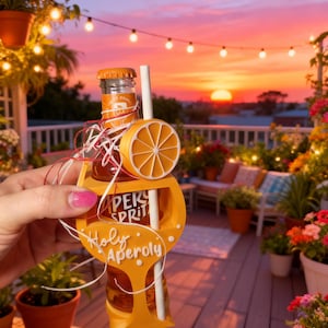 Könnte beinhalten: Eine kleine Glasflasche Aperol Spritz in einem orangefarbenen Halter mit den Worten "Holy Aperoly". Der Halter hat eine dekorative Orangenscheibe und einen Strohhalm. Der Hintergrund zeigt einen Sonnenuntergang und eine Außenumgebung.