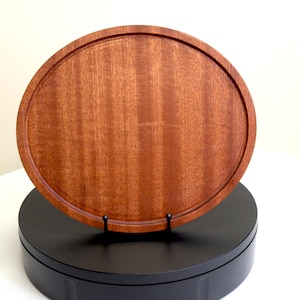 May include: A round, wooden serving tray with a raised edge. The tray is made of reddish-brown wood and sits on a black stand. The tray's diameter is approximately 30 cm.