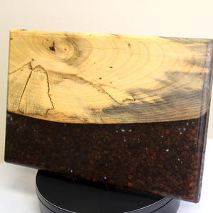May include: A rectangular wooden serving board with a unique design. The top half features light-colored wood with visible grain, while the bottom half is filled with dark brown coffee beans encased in resin. A decorative piece for the kitchen.