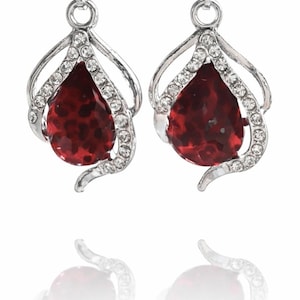 May include: A pair of silver drop earrings. Each earring has a teardrop-shaped, deep red gemstone, encircled by small, clear crystals. The earrings have an openwork design above the gemstone. The earrings are a classic jewelry item.