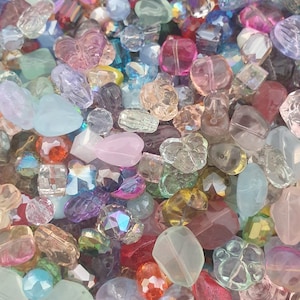 May include: A close-up of a collection of colourful beads in various shapes and sizes. The beads are in shades of pink, purple, blue, and clear, with some iridescent and faceted. The beads are scattered, creating a textured surface.