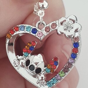 May include: Silver-toned heart-shaped pendant with a cartoon character and colorful gemstones. The pendant features a character with black eyes and colorful accents. A flower with a gemstone center is on the top right of the heart.