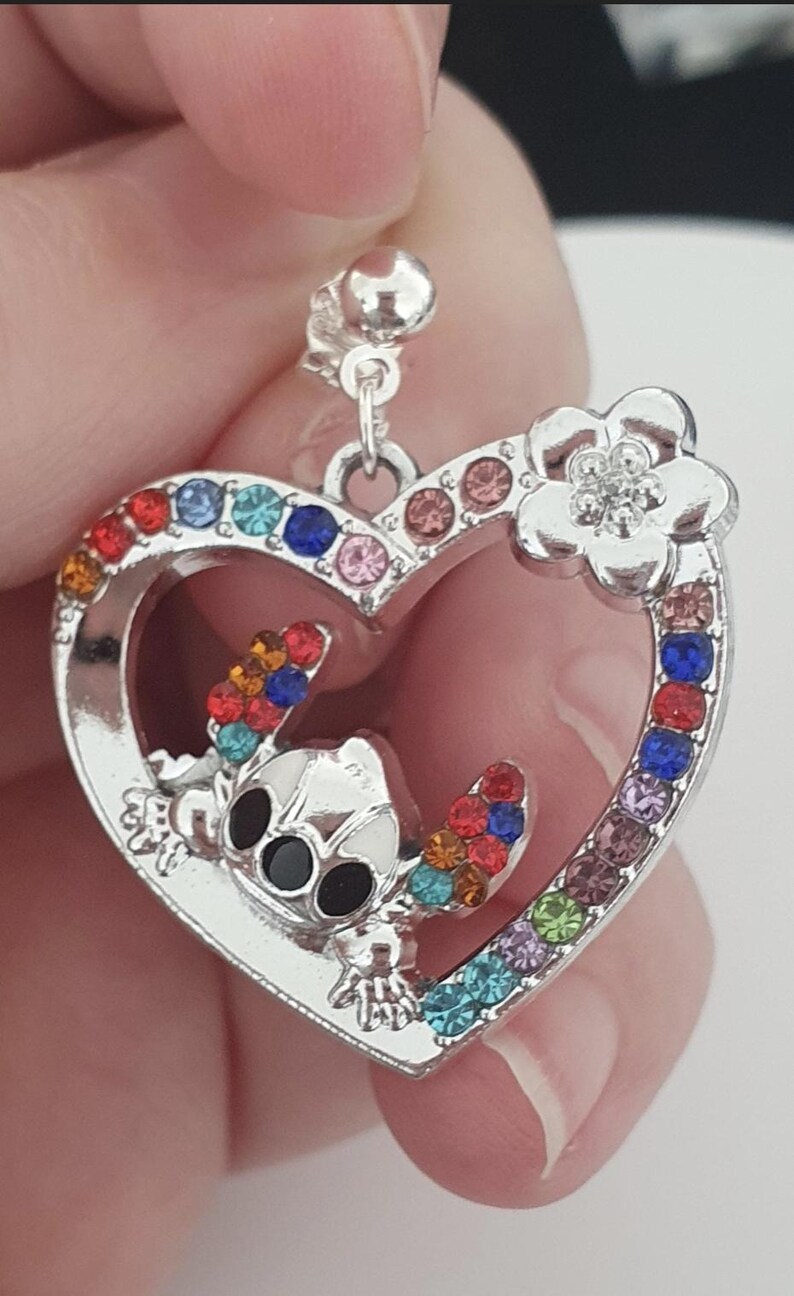 May include: Silver-toned heart-shaped pendant with a cartoon character and colorful gemstones. The pendant features a character with black eyes and colorful accents. A flower with a gemstone center is on the top right of the heart.