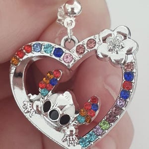 May include: Silver-toned heart-shaped pendant with a cartoon character and colorful gemstones. The pendant features a character with black eyes and colorful accents, surrounded by a border of various colored gemstones. A flower is at the top.