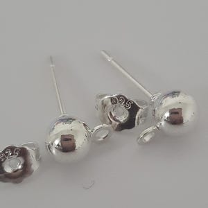 May include: Close-up of several silver ball stud earrings with post and butterfly backings. The earrings are made of shiny, polished metal. The backings are stamped with "925".