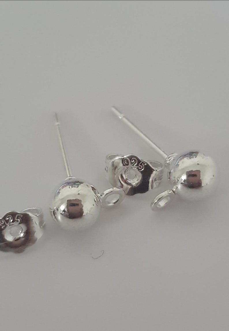 May include: Close-up of several silver ball stud earrings with post and butterfly backings. The earrings are made of shiny, polished metal. The backings are stamped with "925".