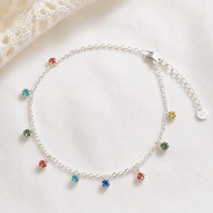 Könnte beinhalten: Ein filigranes silbernes Fußkettchen mit einer Kette, die mit bunten, runden Edelsteinen besetzt ist. Die Edelsteine sind in verschiedenen Farben wie Rot, Blau, Grün und Gelb gehalten. Das Fußkettchen liegt auf einem weißen Hintergrund.