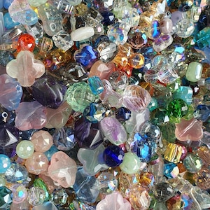 May include: A large collection of loose glass beads in various shapes, sizes, and colours including clear, blue, pink, green, purple, and amber. The beads feature faceted, smooth, and floral-like surfaces with iridescent and reflective finishes.