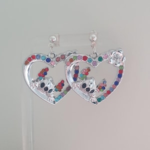 May include: Silver-toned heart-shaped earrings with colorful gemstone accents. Each earring features a cartoon character design and a flower detail. The heart outlines are studded with various colored gems, creating a vibrant and eye-catching accessory.