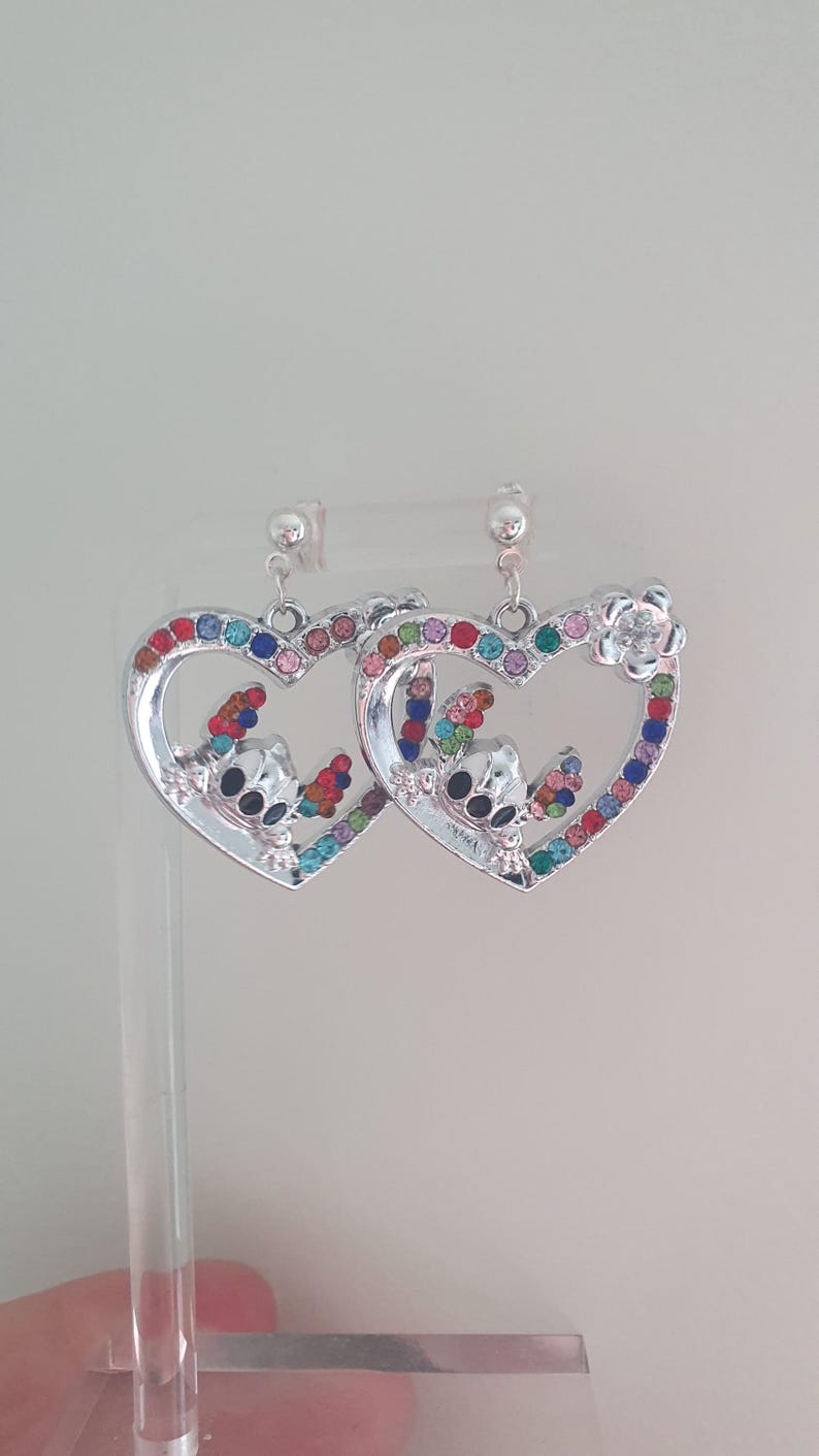 May include: Silver-toned heart-shaped earrings with colorful gemstone accents. Each earring features a cartoon character design and a flower detail. The heart outlines are studded with various colored gems, creating a vibrant and eye-catching accessory.