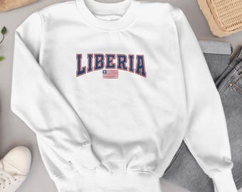 Op de afbeelding: Witte crewneck sweatshirt met het woord "LIBERIA" in marineblauwe gebogen letters met een rode omlijning. Onder het woord staat een kleine Liberiaanse vlag. De sweatshirt is een casual stijl.