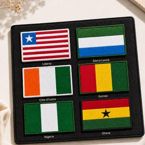 May include: A black rectangular display board featuring six embroidered flag patches with labels underneath. The flags include Liberia, Sierra Leone, Côte d'Ivoire, Guinea, Nigeria, and Ghana, each rendered in their respective national colors and designs.