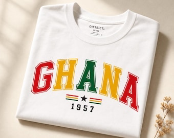 Op de afbeelding: Een wit T-shirt met korte mouwen waarop het woord GHANA staat in een college-lettertype met rode, gele en groene letters. Onder de tekst staat een kleine zwarte ster gecentreerd tussen twee horizontale rode, gele en groene strepen, met het jaartal 1957 in het zwart.