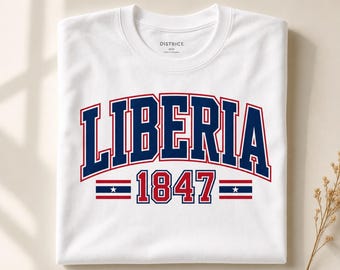 Op de afbeelding: Een wit T-shirt met korte mouwen waarop het woord LIBERIA staat in een grote, gebogen marineblauwe lettertype met een rode omlijning. Onder de tekst staat het getal 1847 in het rood met een marineblauwe omlijning, geflankeerd door twee strepen.