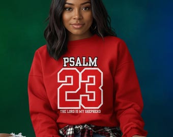 Op de afbeelding: Een rode sweatshirt met de tekst "PSALM 23" in witte blokletters. Onder het nummer 23 staat de tekst "THE LORD IS MY SHEPHERD". De persoon draagt een blauwe spijkerbroek en een geruit overhemd om de taille geknoopt.
