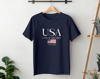 Op de afbeelding: Marineblauw t-shirt met het woord "USA" in wit, erboven "LAND OF THE FREE" en een kleine Amerikaanse vlag. Onder de vlag staat "-EST. 1776-". Het t-shirt hangt aan een houten hanger.