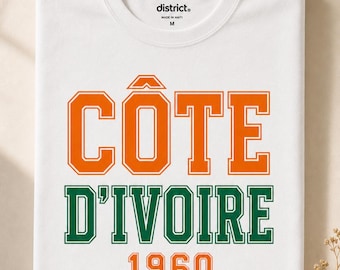Op de afbeelding: Een wit T-shirt met korte mouwen voorzien van de tekst CÔTE D'IVOIRE in oranje en groene blokletters, met het jaartal 1960 in het oranje eronder gedrukt. Het shirt heeft een ronde hals en een district merklabel aan de binnenkant van de kraag.