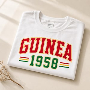 以下が含まれることがあります： 黄色い縁取りの赤い大きな文字でGUINEAと書かれ、その下に黄色い縁取りの緑色の数字1958が配置された白い半袖Tシャツ。両サイドには赤、黄、緑の水平なストライプがあしらわれています。