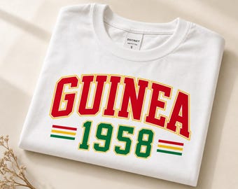 Op de afbeelding: Een wit t-shirt met korte mouwen met het woord GUINEA in grote rode letters met een gele omlijning, gevolgd door het getal 1958 in het groen met een gele omlijning, geflankeerd door horizontale rode, gele en groene strepen aan beide kanten.