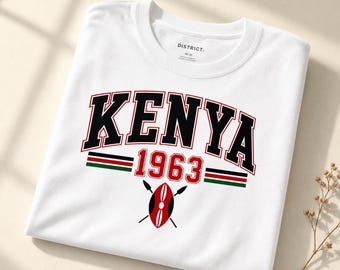 Op de afbeelding: Een wit t-shirt met ronde hals voorzien van het woord KENYA in grote zwarte letters met rode omlijning, het getal 1963 in het rood en een afbeelding van een traditioneel schild met twee gekruiste speren. Het ontwerp bevat horizontale rode, zwarte en groene strepen.